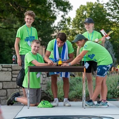 Five summer camp teens wearing bright green t-shirts stand behind a small table. a