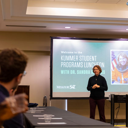 Speaker presenting at a student programs luncheon in a conference room with attendees seated at tables.