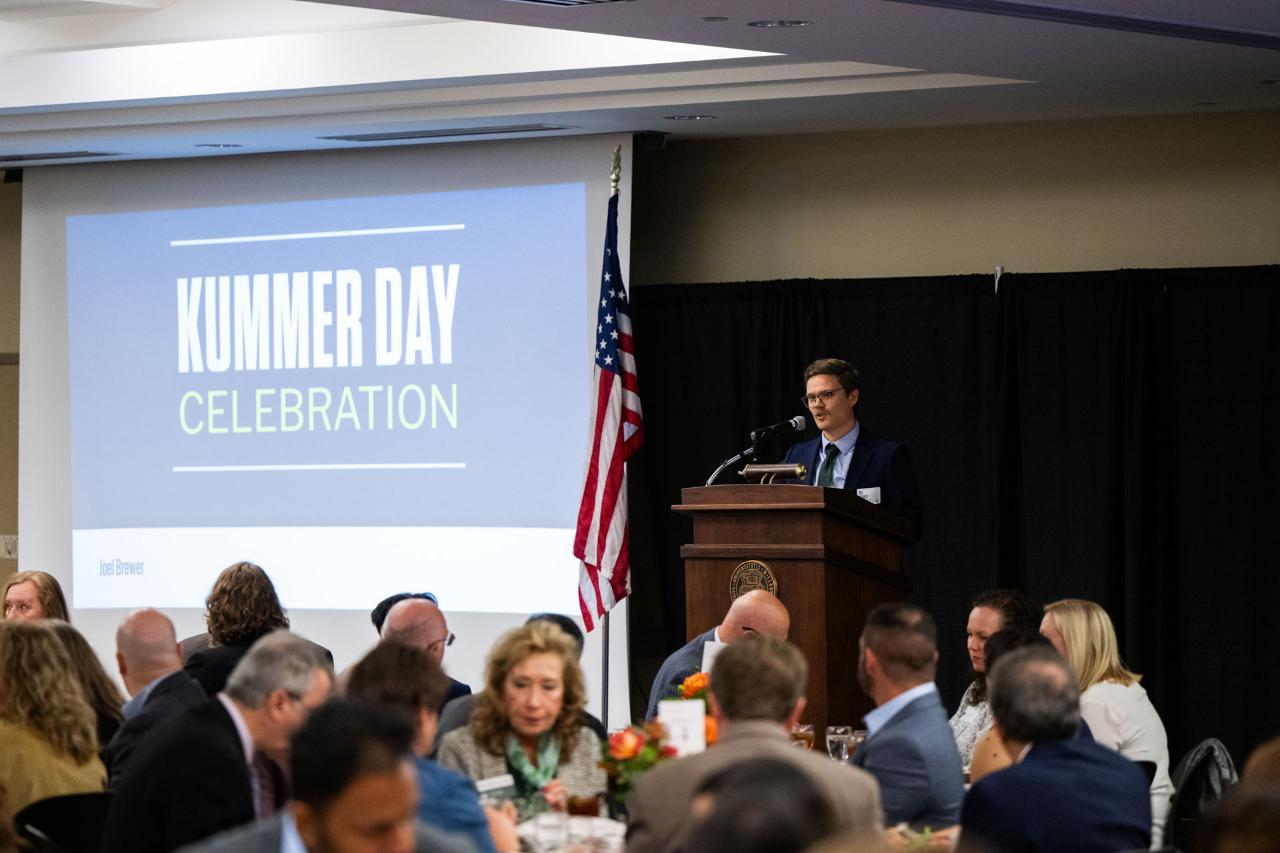 Man standing at a podium speaking to people siting at round tables for lunch.