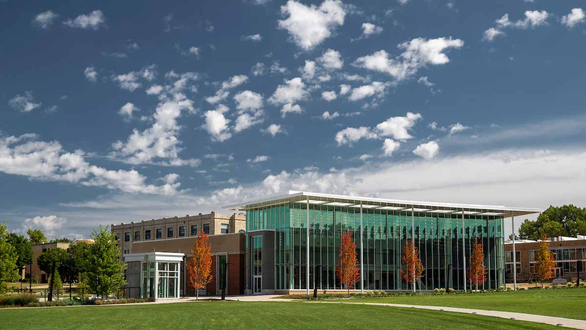 Glass-fronted building on campus with a green lawn and trees under a blue sky with scattered white clouds.