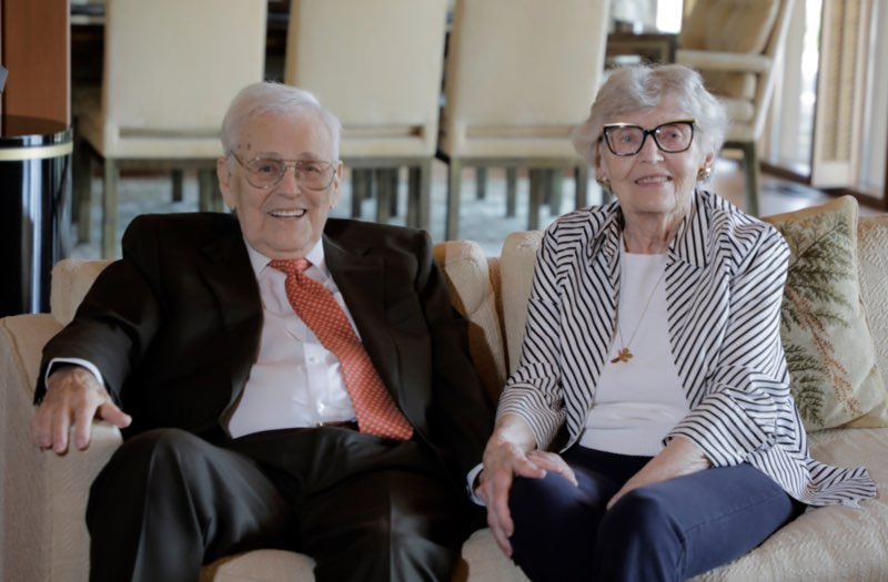 An elderly man and woman sit side by side on a couch, smiling warmly at the camera.